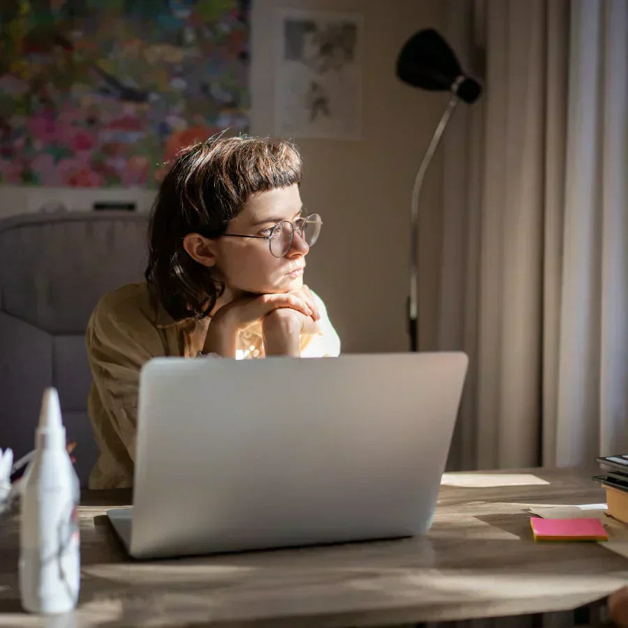 Une jeune femme pensive aux lunettes regarde au loin, assise devant un ordinateur portable sur un bureau en bois