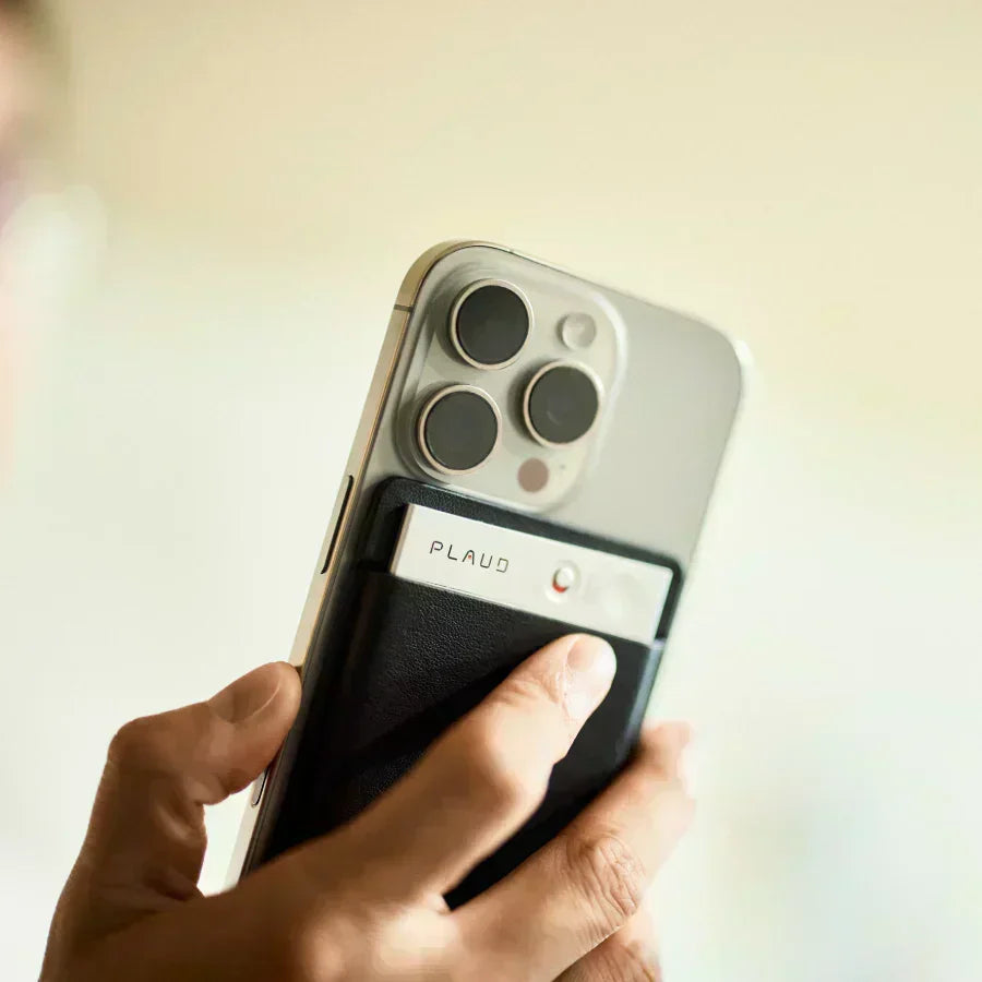 Une main tient un smartphone argenté avec un Plaud Note noir et argenté fixé magnétiquement à l'arrière.