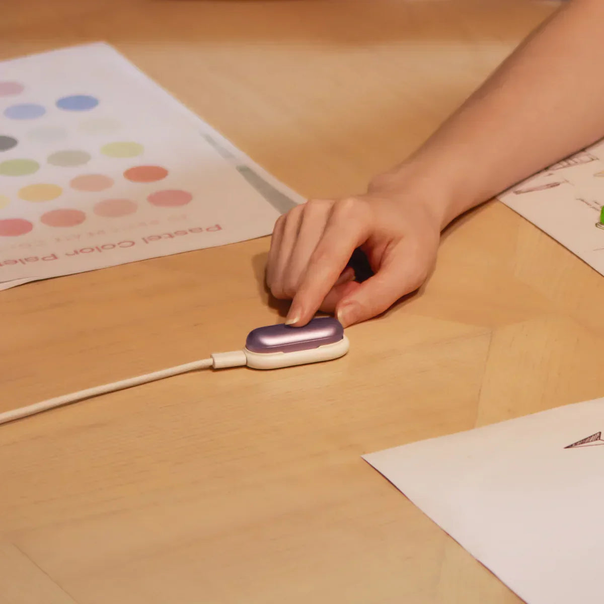 Une main touche un Plaud NotePin violet et blanc, branché à un câble de charge sur une table en bois clair