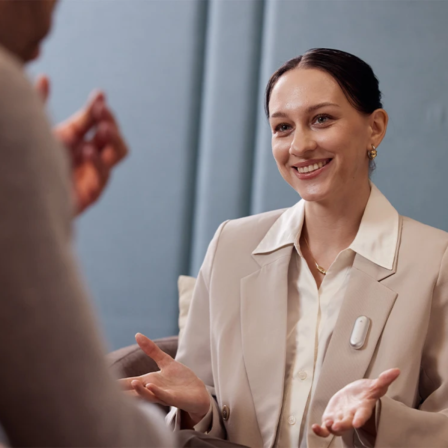 Une femme souriante discute, son Plaud NotePin blanc accroché à son blazer, lors d'une conversation professionnelle.