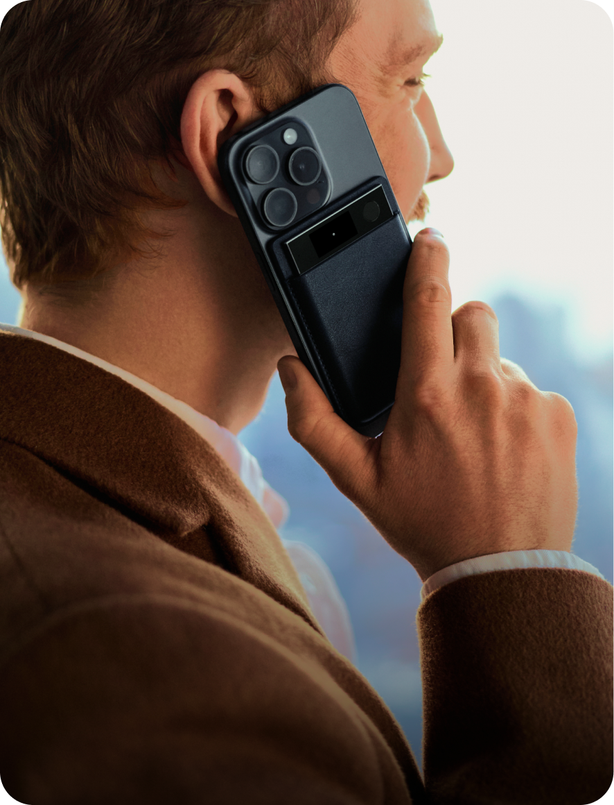 Un homme portant une veste marron tient un smartphone noir contre son oreille, avec un Plaud Note Pro fixé au dos de l'appareil.