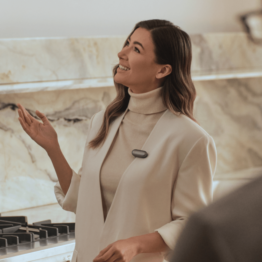Une femme souriante, vêtue d'une veste claire, porte un Plaud NotePin S gris foncé sur sa poitrine dans une cuisine moderne.