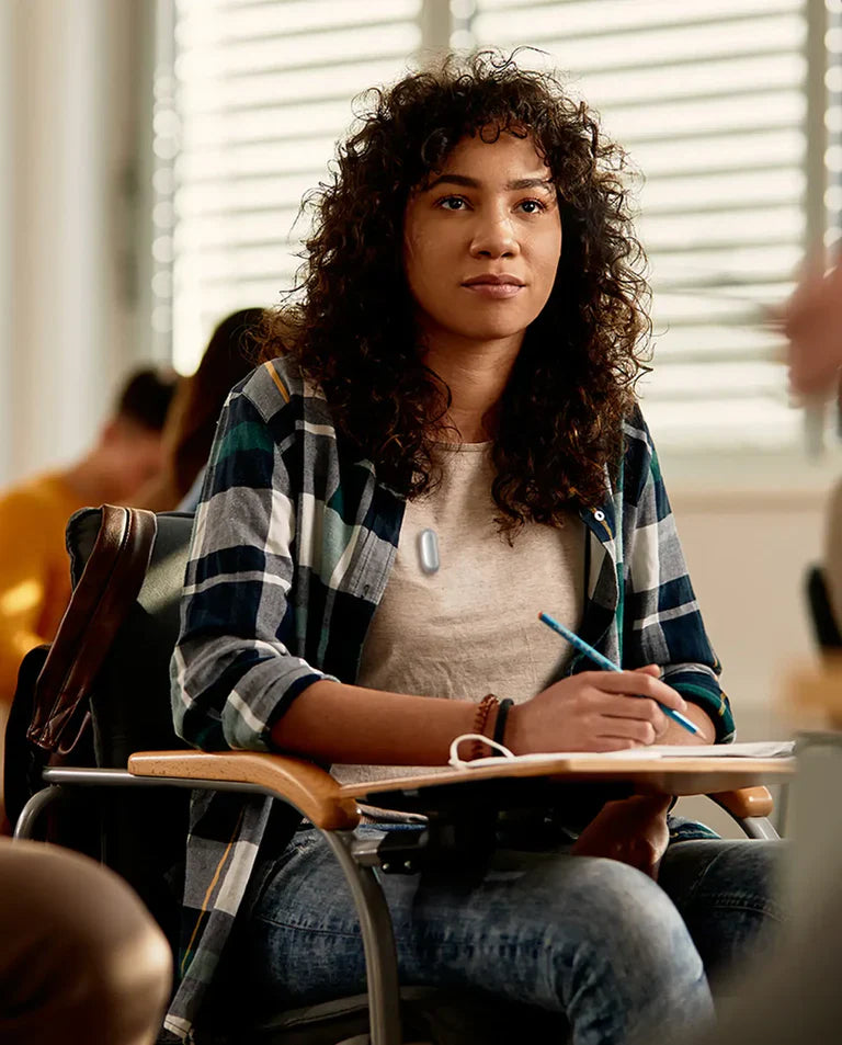Une jeune femme attentive en classe porte un Plaud NotePin gris clair sur son t-shirt beige, prenant des notes.