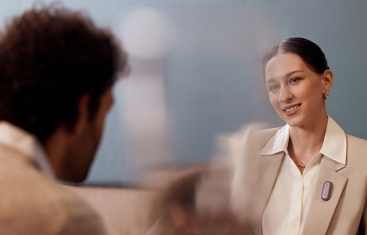 Une femme souriante porte un Plaud NotePin S rose doré sur son blazer lors d'une conversation professionnelle.
