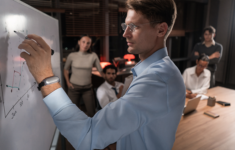 Un homme en chemise bleue présente des graphiques sur un tableau blanc lors d'une réunion professionnelle