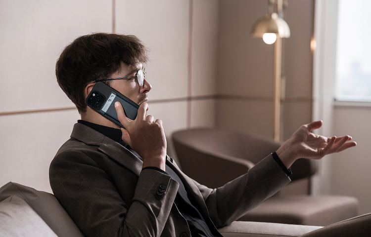 Un homme en costume parle au téléphone, un Plaud Note Pro noir fixé à son smartphone, gesticulant de la main dans un salon.