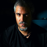 Portrait d'un homme aux cheveux et à la barbe grisonnants, regardant intensément la caméra sur un fond sombre.