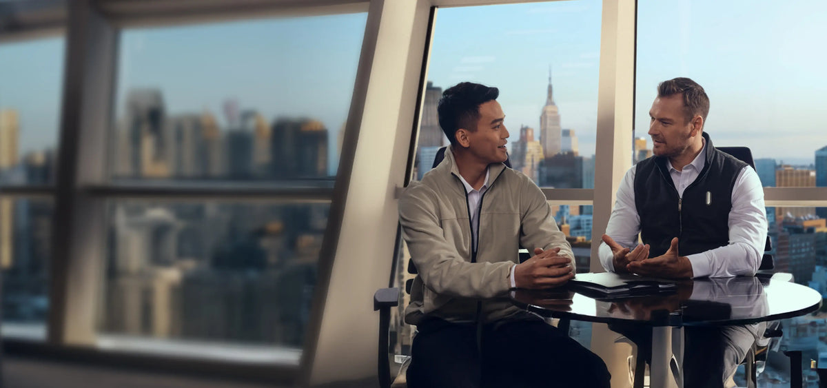 Deux hommes en conversation dans un bureau moderne avec vue sur la ville, un enregistreur vocal Plaud AI noir sur la table.