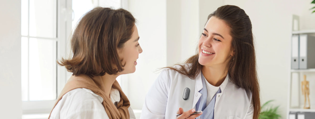 Une femme médecin souriante en blouse blanche discute avec une patiente, portant un enregistreur portable Plaud NotePin S gris sur sa blouse.