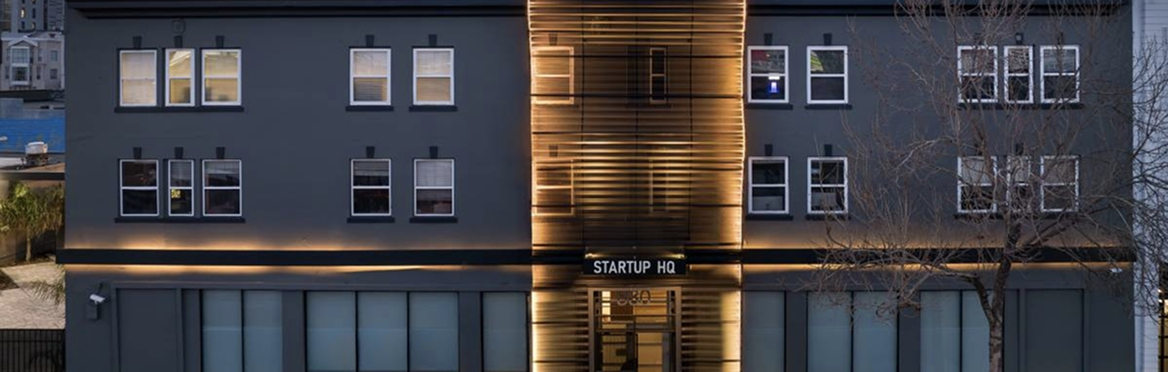 Façade d'un bâtiment moderne gris foncé éclairé par des bandes lumineuses chaudes, avec l'inscription 'STARTUP HQ'.