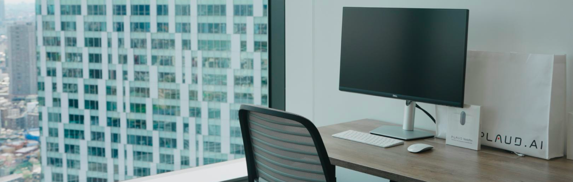 Bureau moderne avec écran, clavier, souris, un sac Plaud et un paquet Plaud Note sur une table, vue sur la ville.