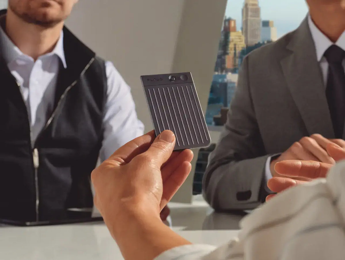 Une main présente un Plaud Note gris foncé à deux hommes en tenue de travail, dans un bureau moderne.