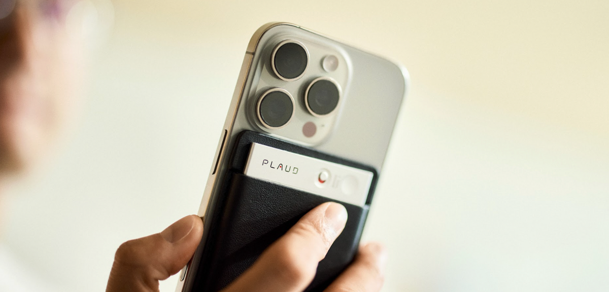 Une main tient un smartphone argenté avec un Plaud Note argenté et son point rouge, fixé à l'arrière dans un étui noir.